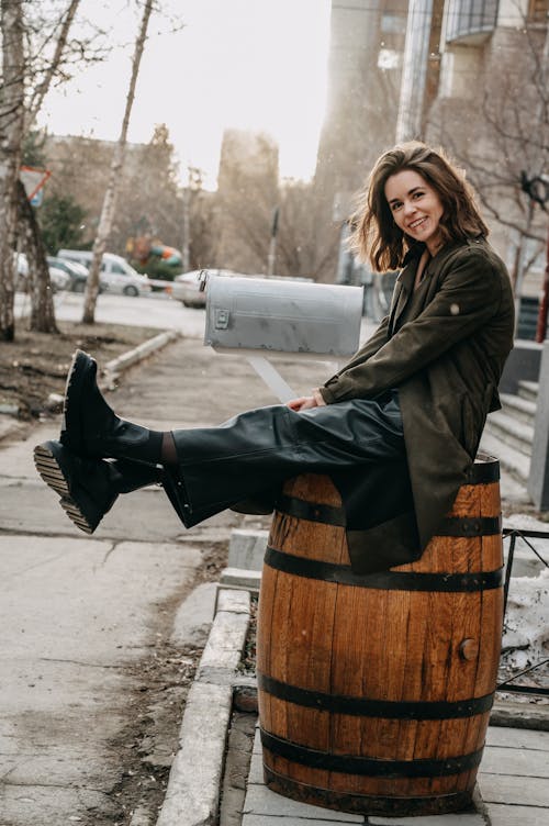 Full body of woman in warm clothes looking at camera and smiling while sitting on wooden barrel in town