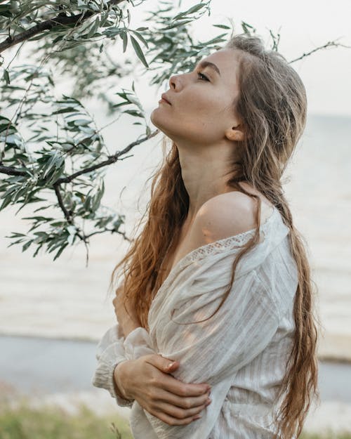 Side view of female with long hair standing near green plants and raising head against blurred sea