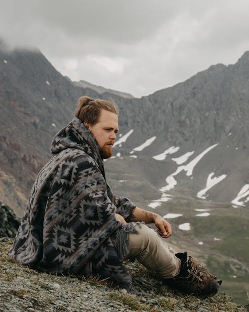 Side view of male traveler in poncho sitting on hilltop while contemplating majestic mountain in foggy day