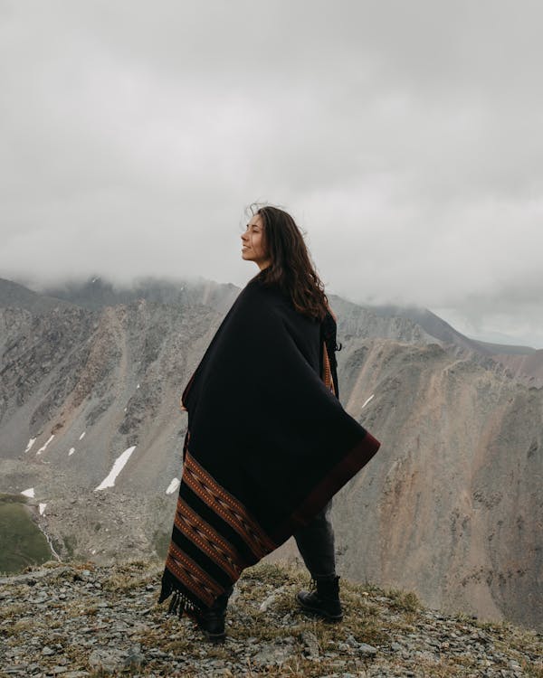 Ethnic female in warm cloak standing on edge of precipice in mountains and looking away in cloudy day