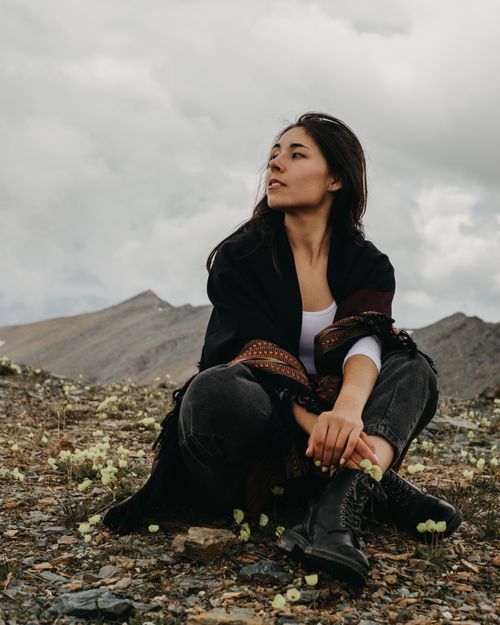 Free Full body of ethnic female sitting on ground with crossed legs and looking away against mountains in overcast weather Stock Photo