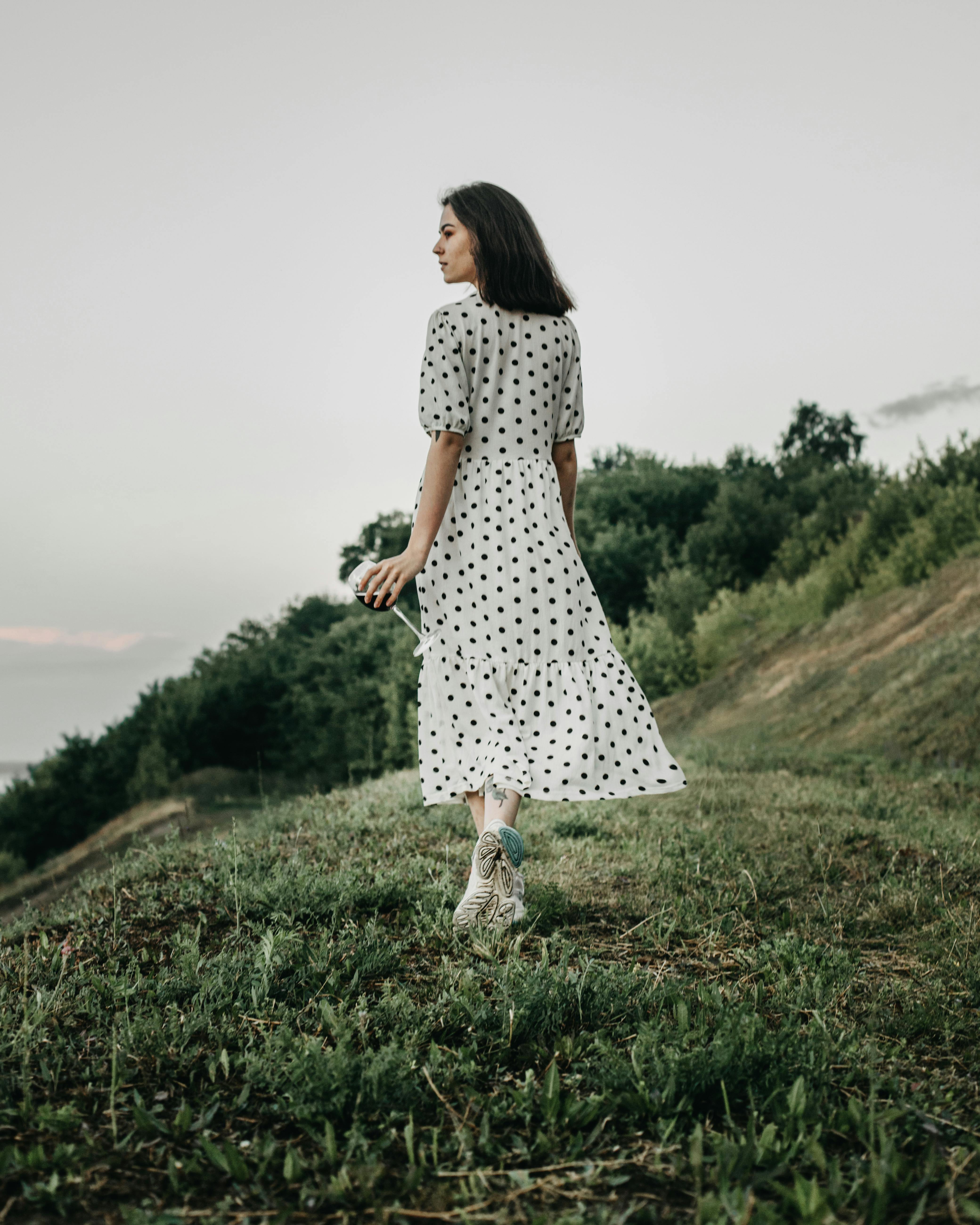 Back view of full body female in polka dot white dress walking on grassy meadow in countryside and looking away