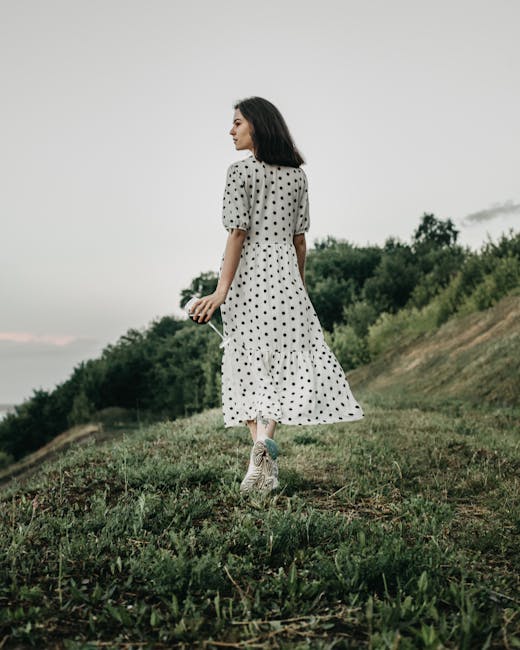 Back view of full body female in polka dot white dress walking on grassy meadow in countryside and looking away