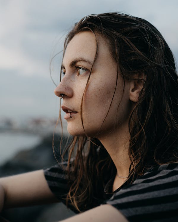 Side view of young dreamy female with lip piercing and wet hair looking away thoughtfully while sitting on shoreline