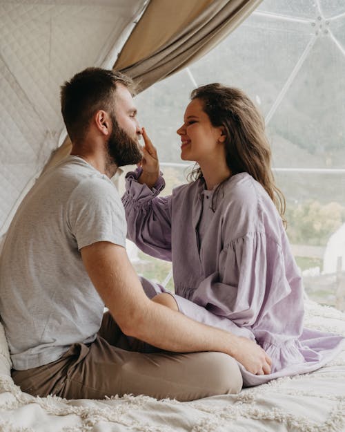 Side view of loving couple sitting on bed and looking at each other while wife touching husband nose