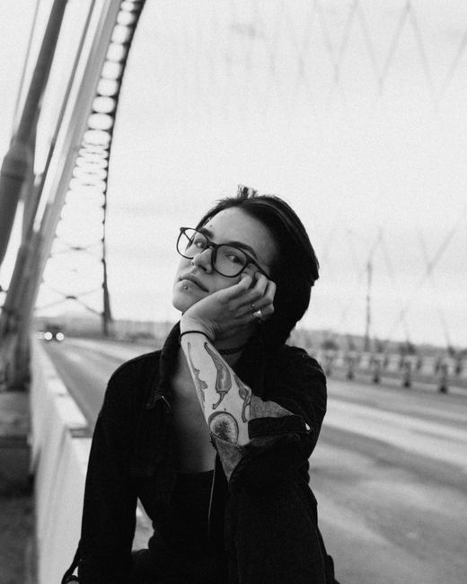 Black and white of extravagant woman leaning on hand while sitting on concrete fence of bridge and looking at camera