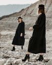 Full body of young alike women in similar black dresses and hats standing opposite each other on stony ground in desert