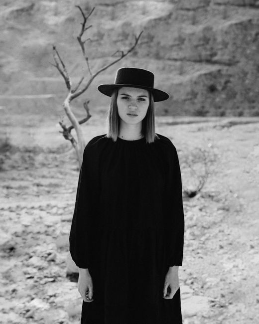 Black and white of young female in black clothes and hat standing in countryside and looking at camera