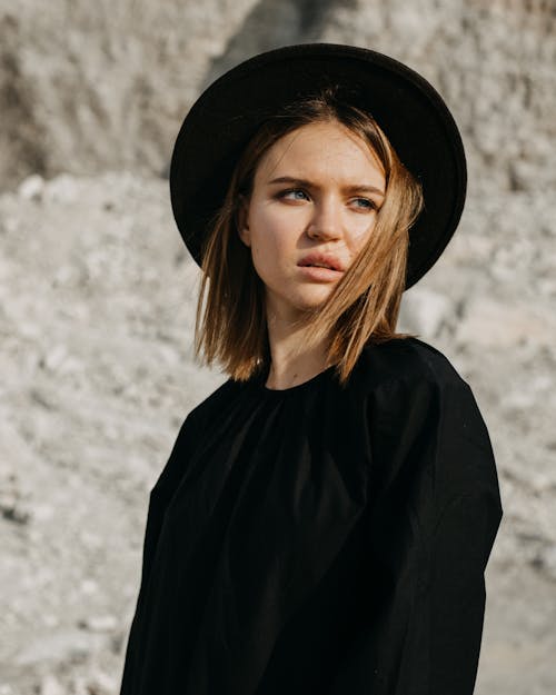 Young trendy female in black outfit and hat standing against gray rocky cliff in sunlight and looking away