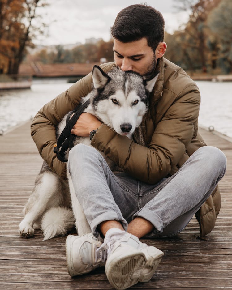 Man Sitting On Wooden Dock While Hugging His Pet