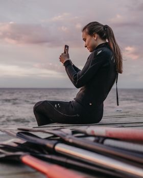 Woman in a wetsuit sits on a dock capturing the ocean view with her smartphone at sunset.