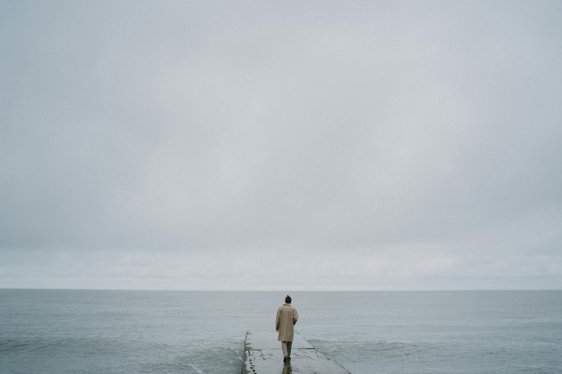 A lone person walks along a concrete pier under a cloudy sky, gazing at the expansive ocean horizon.