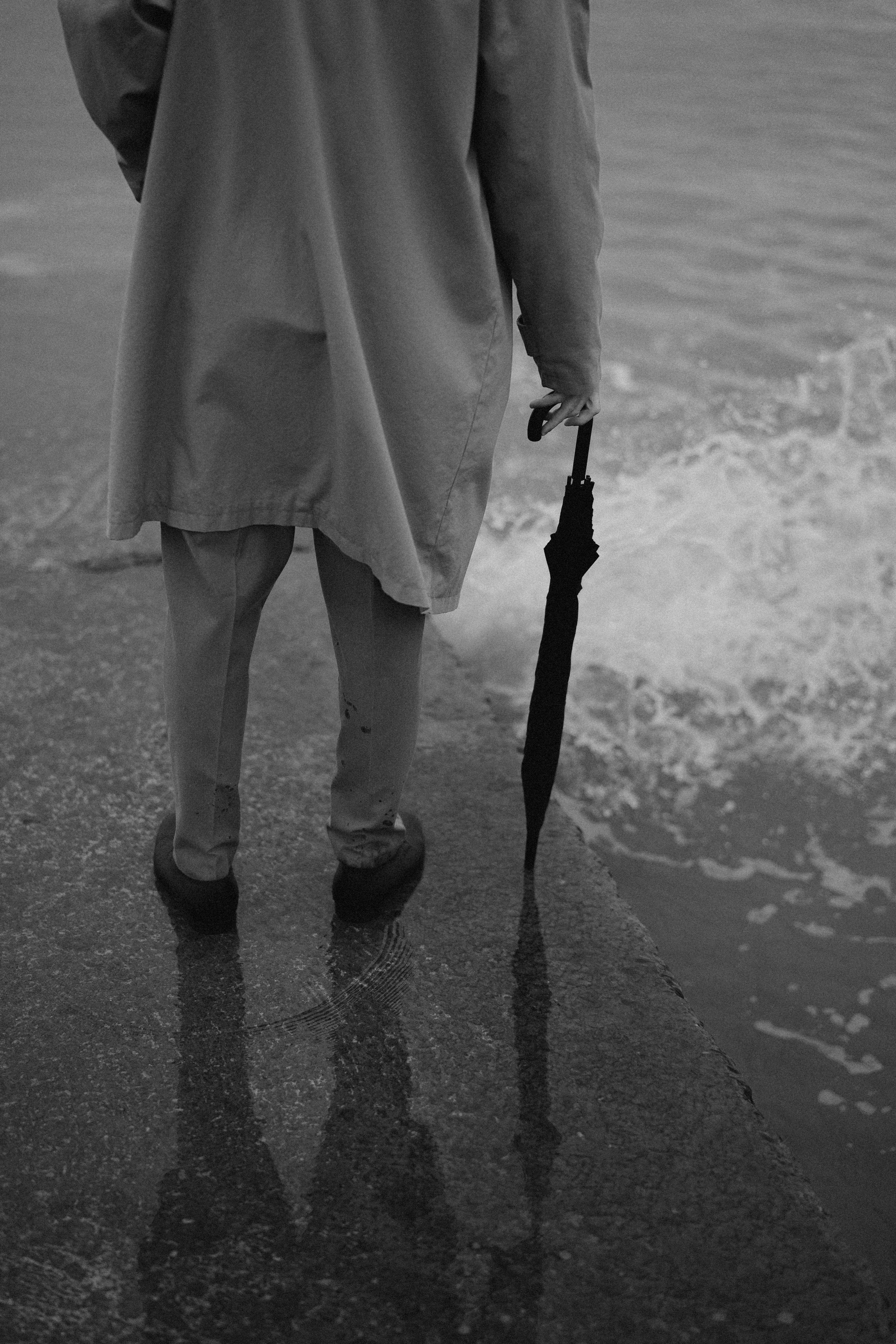 A black and white image of a person holding an umbrella standing by the water's edge.