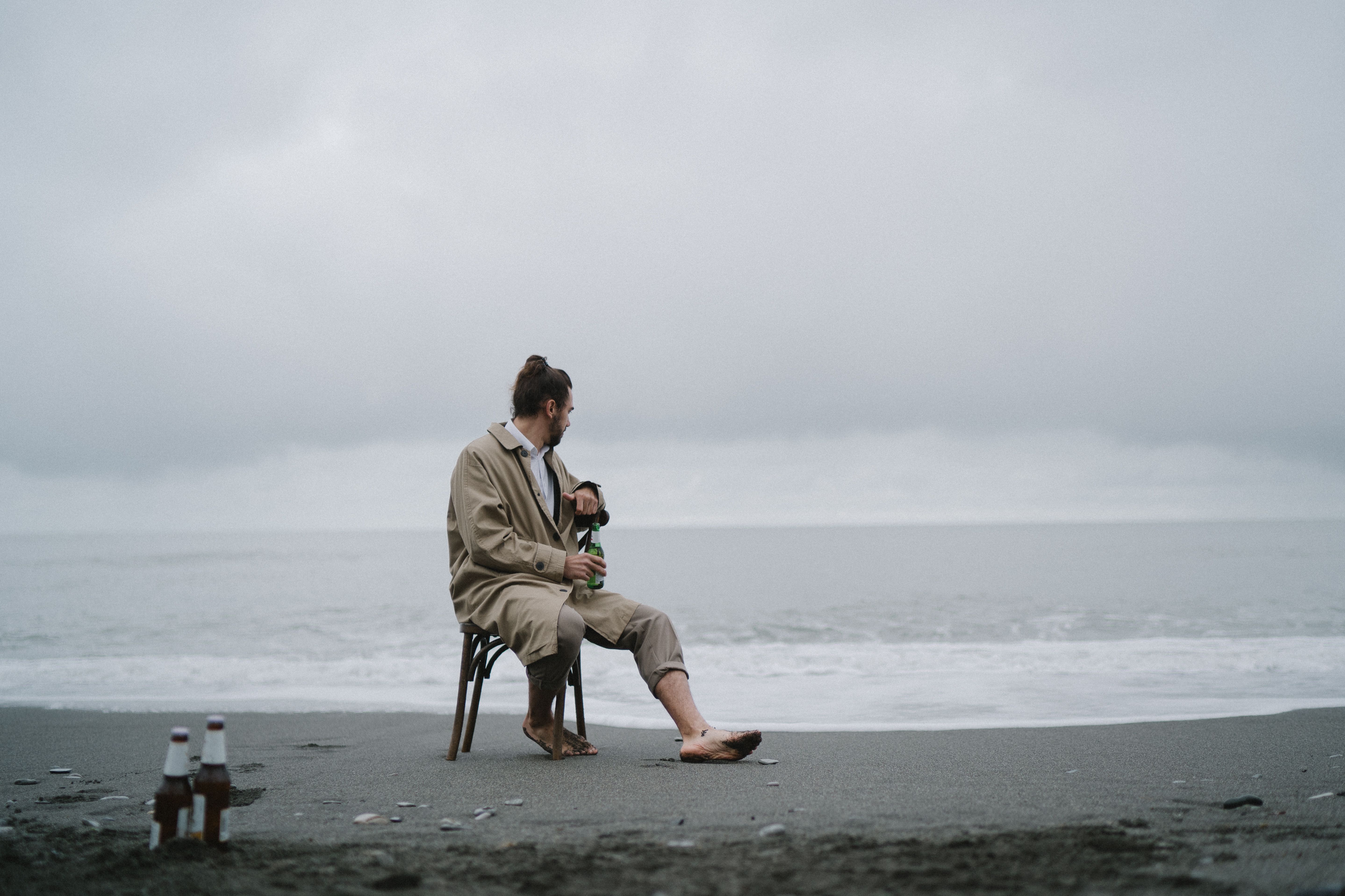 A man sits barefoot on a chair at the beach, with bottles and the sea visible under an overcast sky.