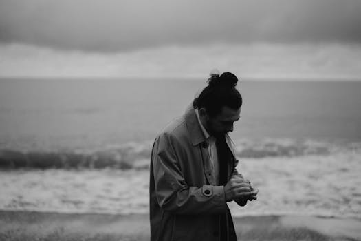 A man with a bun in a coat looks introspectively by the sea in a monochrome setting.