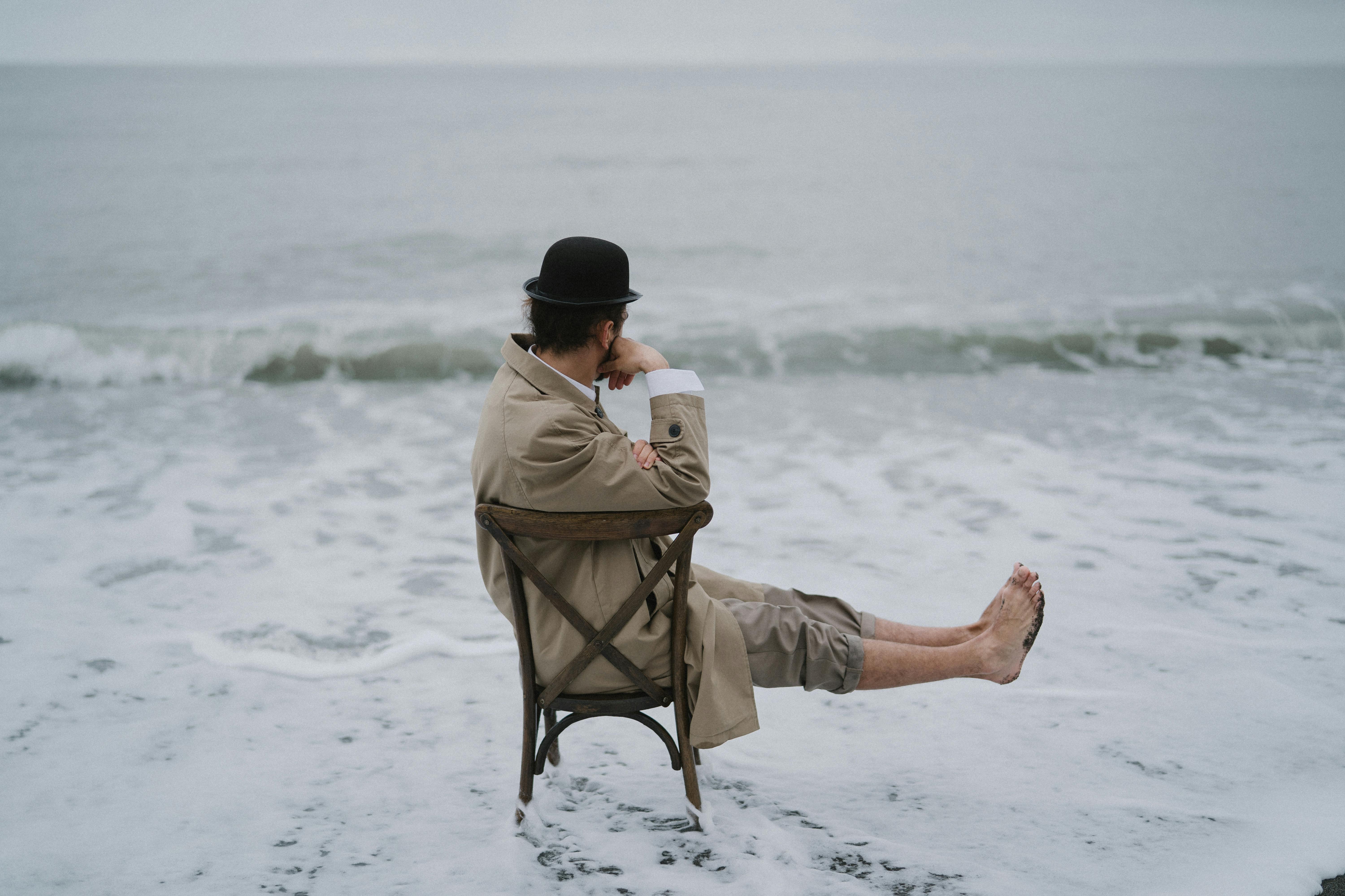 man in brown jacket sitting on brown chair on beach