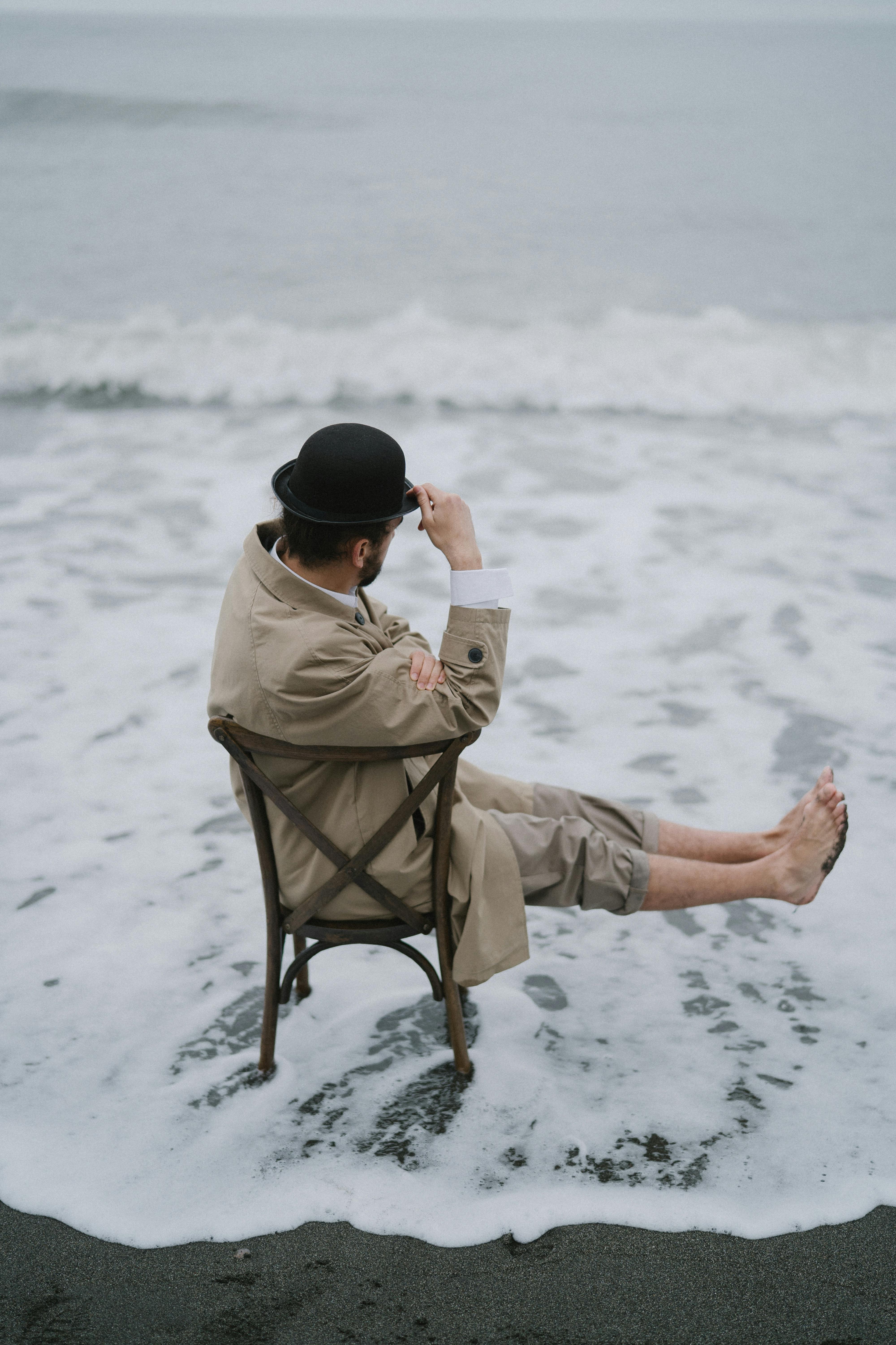 穿着米色外套和圆顶硬礼帽的男人坐在海边的椅子上，拥抱孤独