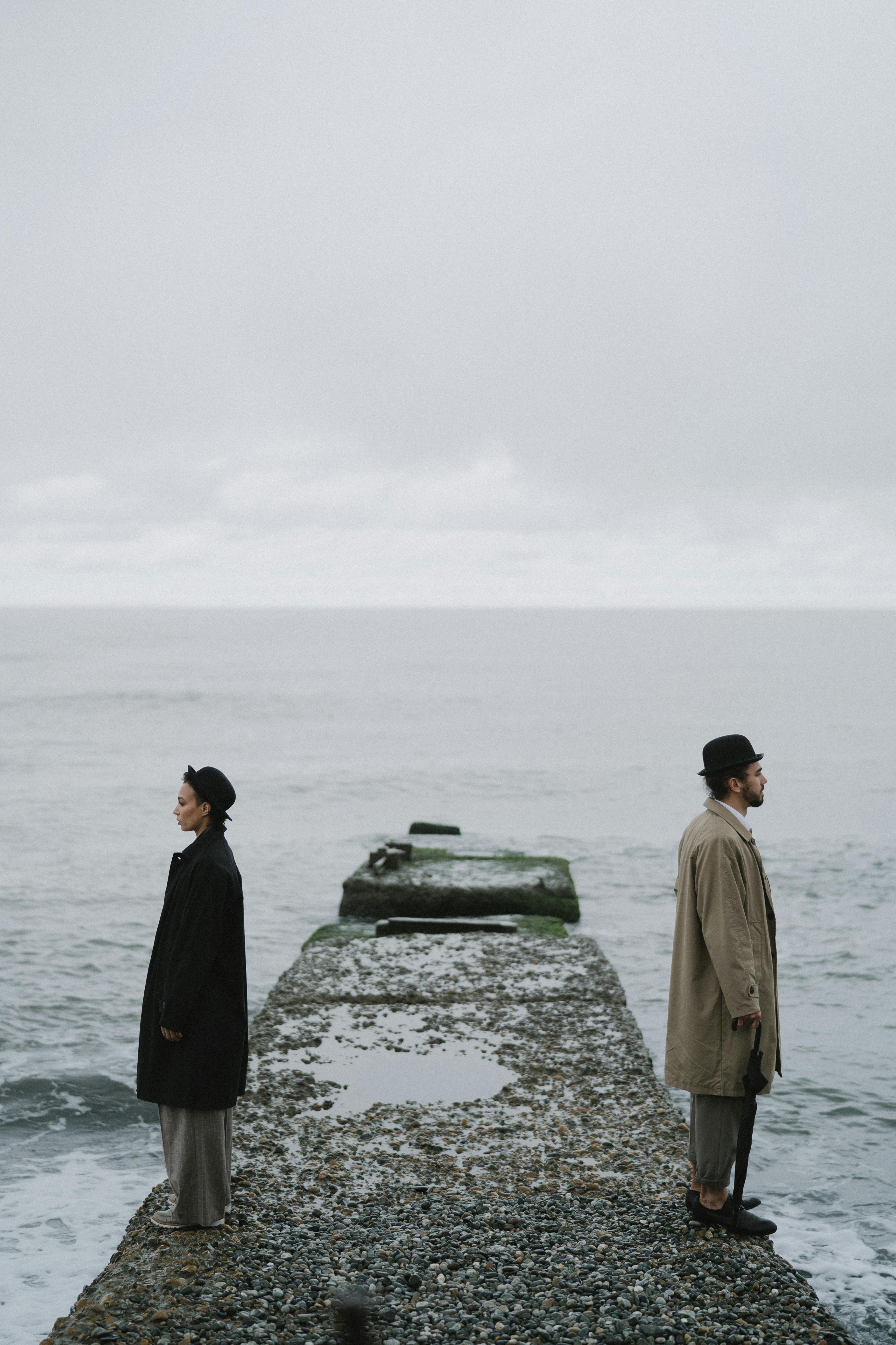 在阴云密布的天空下，两个人背靠背站在海边的码头上