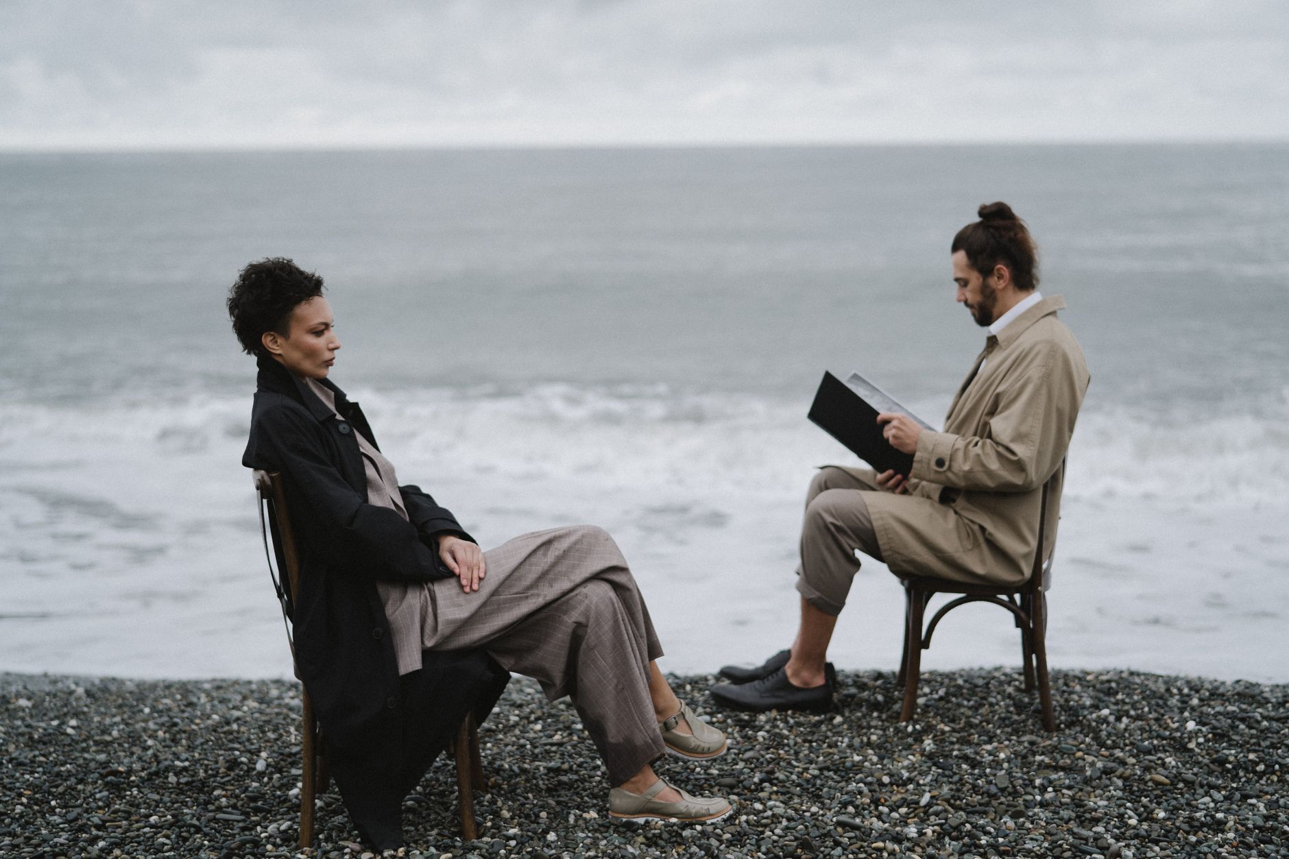 Two adults in conversation on a pebbled beach, calm sea in background.