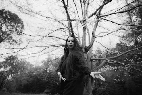 Grayscale Photo of Woman in Black Long Sleeve Dress Standing Near Bare Tree