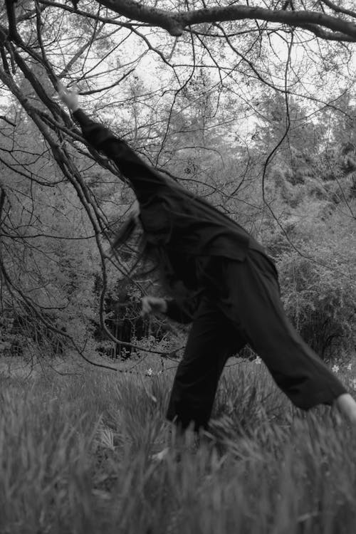 Monochrome Photo of Woman in Black Jacket and Pants dancing near Bare Trees