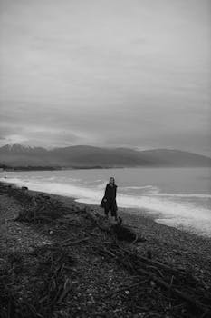 A moody black and white photograph of a person standing on a driftwood-strewn beach.