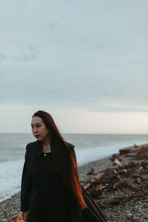 Free Woman in black outfit walking along a rocky coast under an overcast sky, reflecting a moody atmosphere. Stock Photo