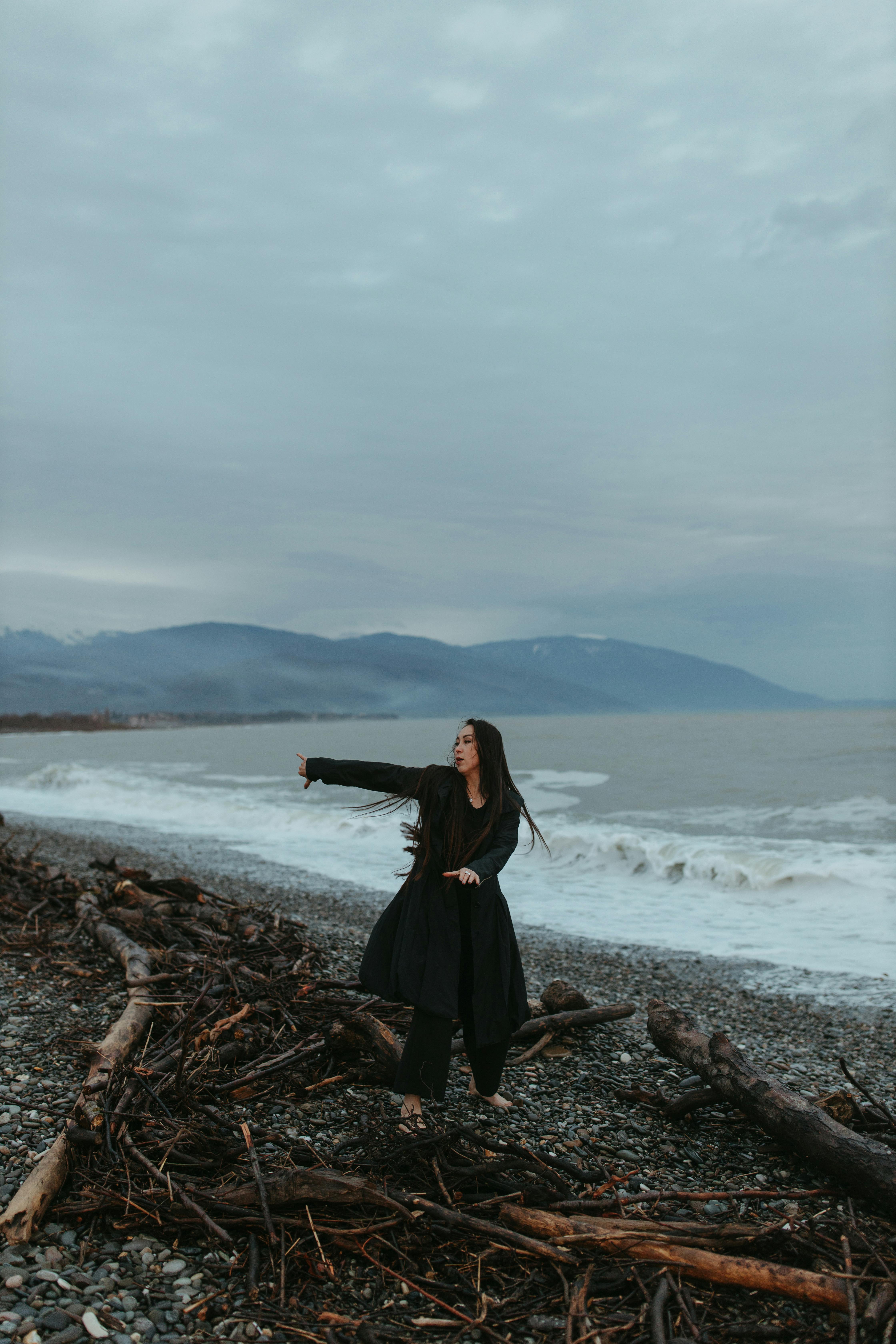 A woman dances gracefully on a pebbled beach with driftwood, under a moody sky.