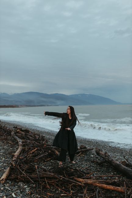 A woman dances gracefully on a pebbled beach with driftwood, under a moody sky.