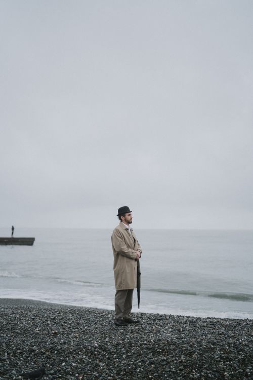 Free Man in brown coat and hat standing alone on foggy seashore. Moody, introspective atmosphere. Stock Photo