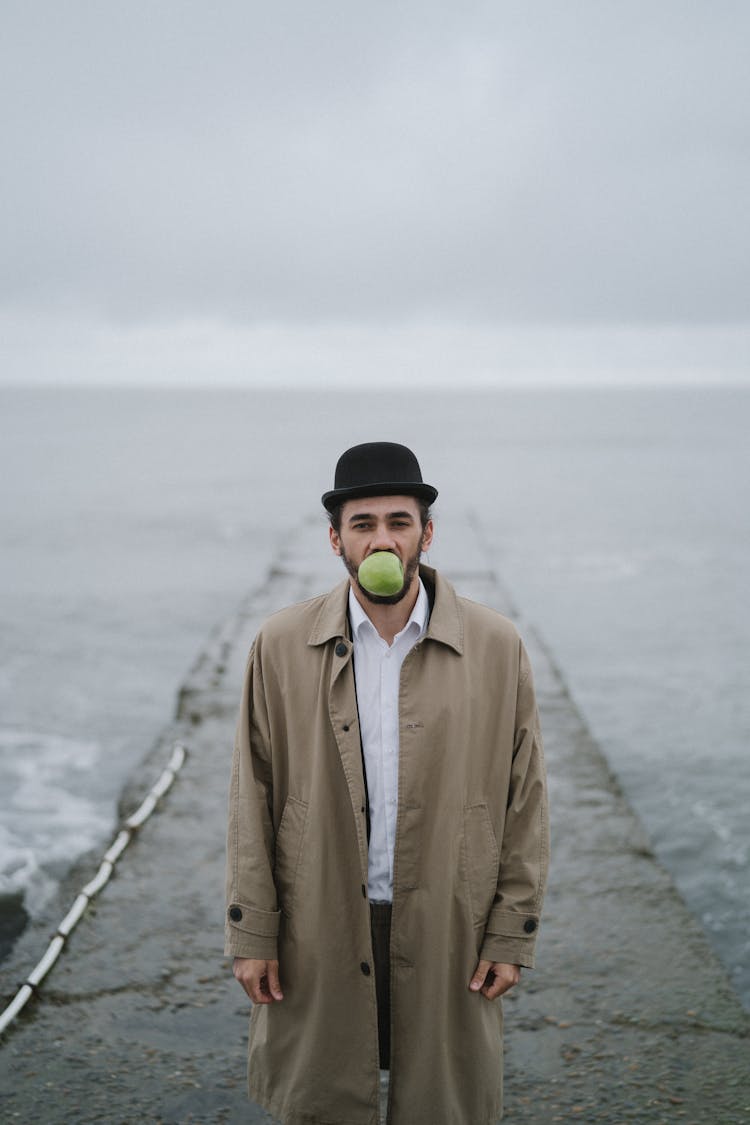 A Man In Brown Coat Standing On A Concrete Dock Near The Sea With Apple On His Mouth