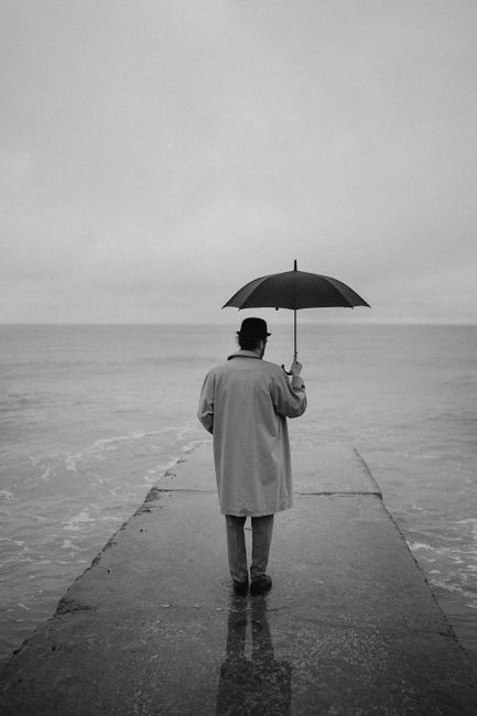 Black and white photo of a person holding an umbrella, standing on a pier, gazing at the sea.