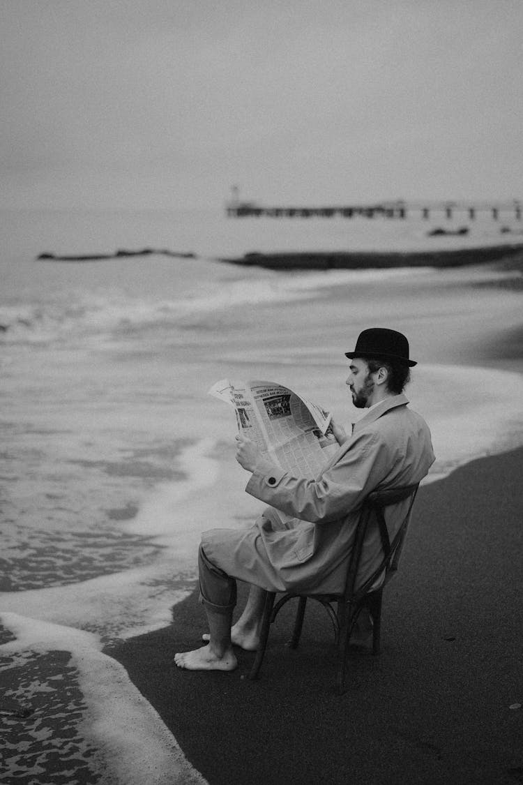 Grayscale Photo Of A Man Reading A Newspaper While Sitting On A Chair At The Beach