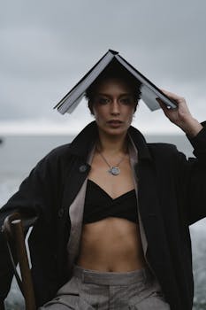 Woman sitting near water, holding a book over her head as a hat, with a moody sky in the background.