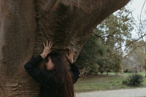 Free A woman in black clothing leans against a large tree trunk, creating a contemplative and serene scene in a lush outdoor setting. Stock Photo