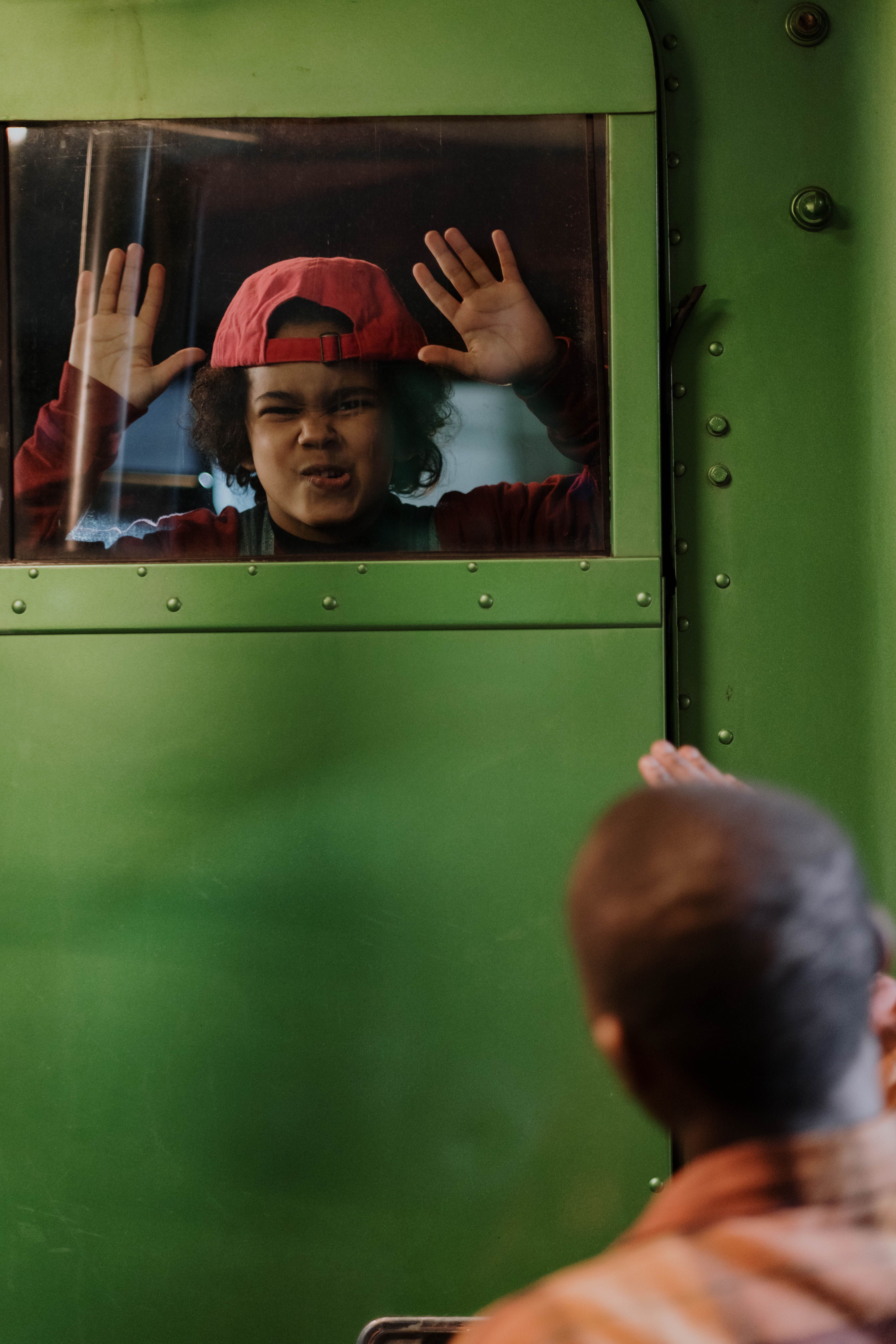 A child wearing a red cap playfully presses face against a green train door window while waving to a person outside.