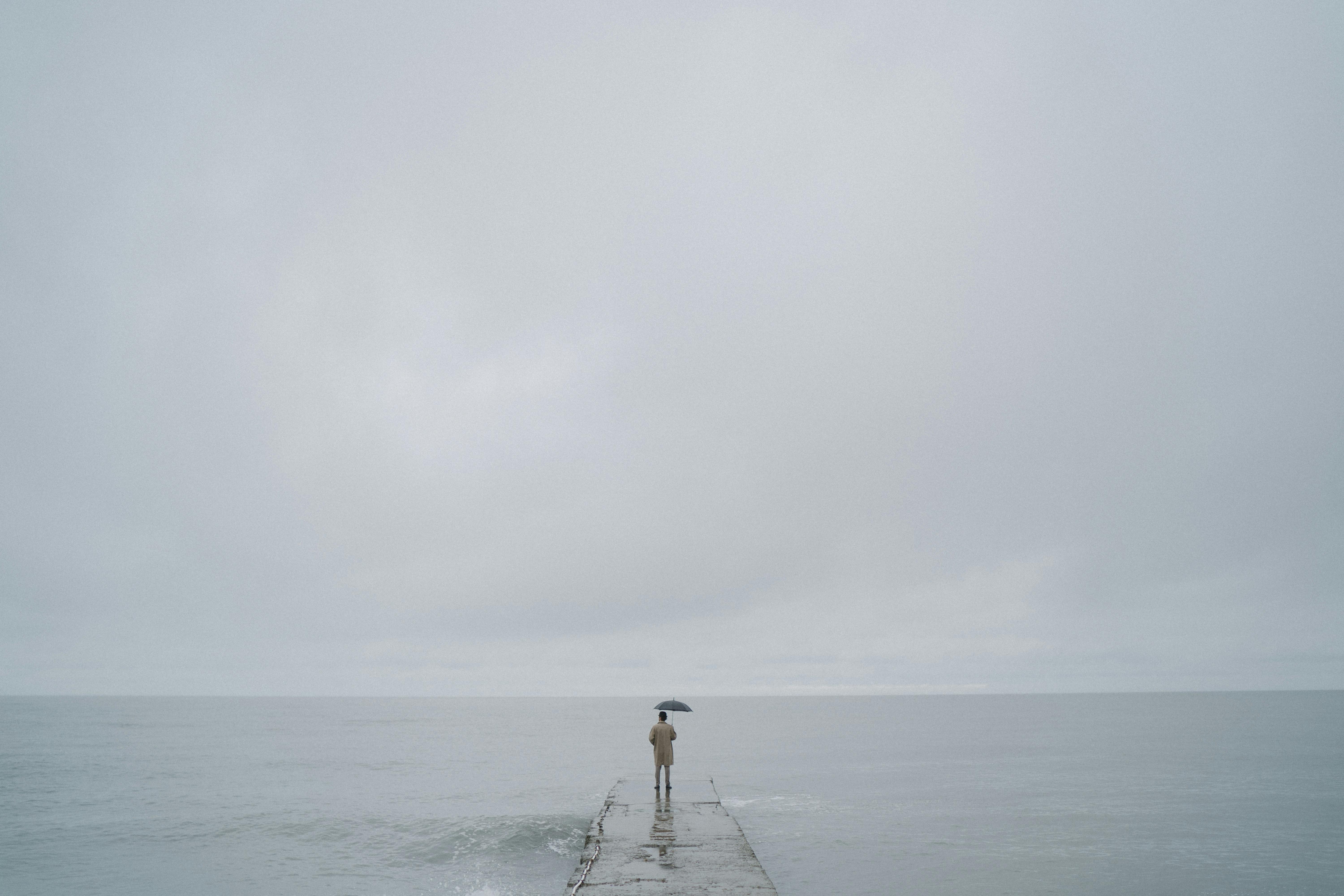 一个带伞的孤独的人站在码头上，在灰色多云的天空下延伸到广阔的大海上