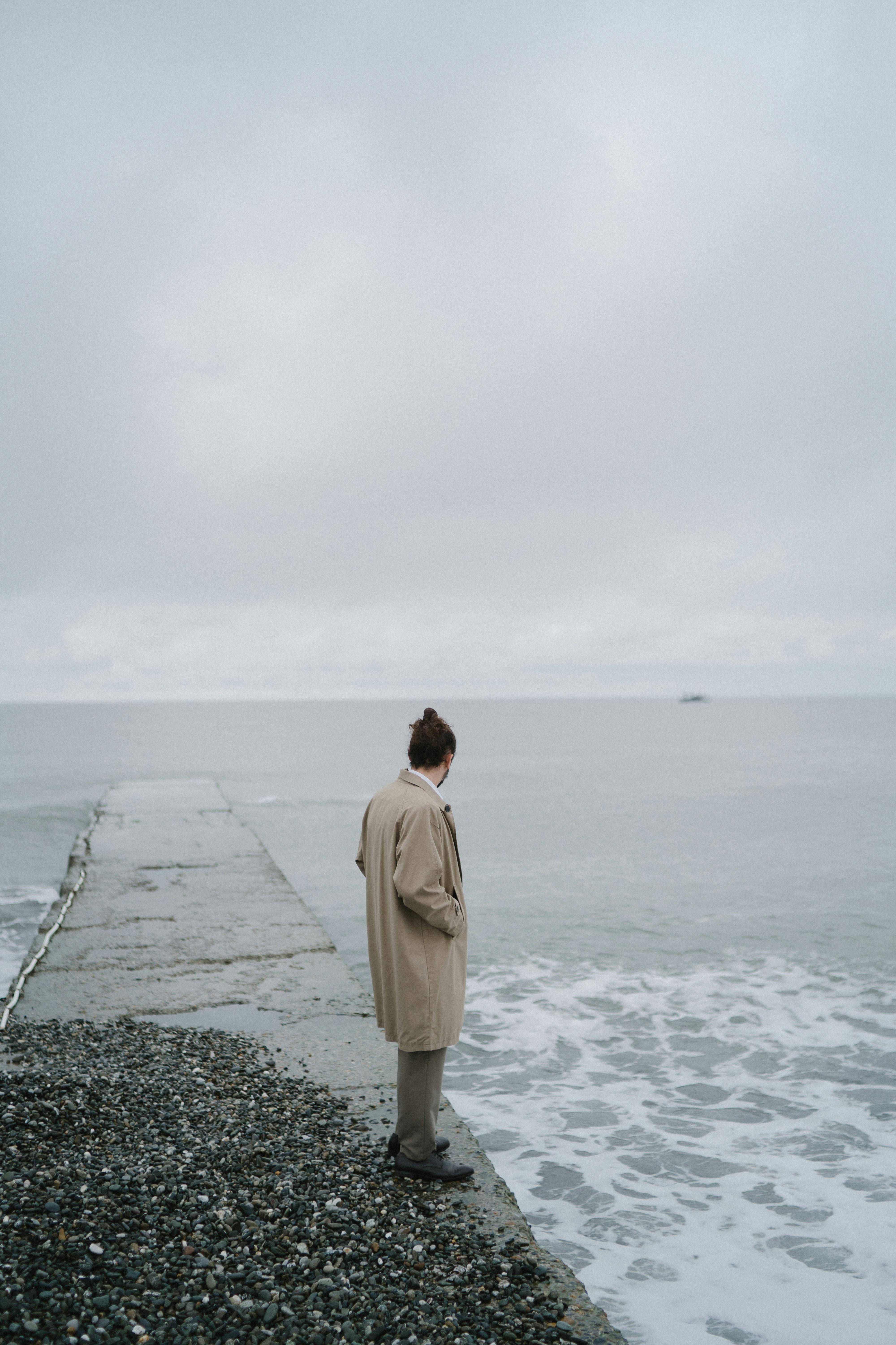 A person stands thoughtfully on a concrete dock by the ocean under a gray sky.