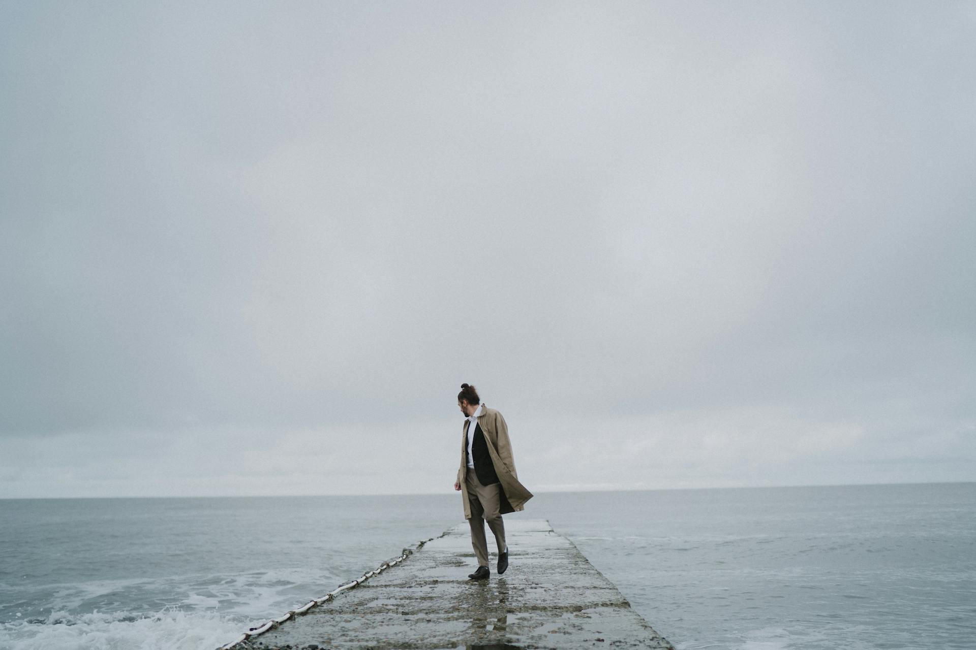 Stylish man walking on a concrete dock by the ocean under a cloudy sky, exuding a fashionable vibe.