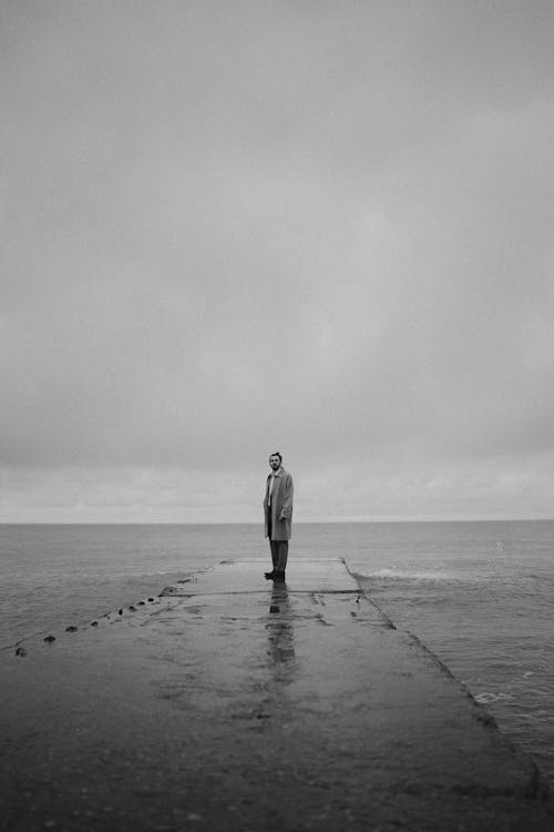 Grayscale Photo of a Person Standing on Concrete Pier under the Sky