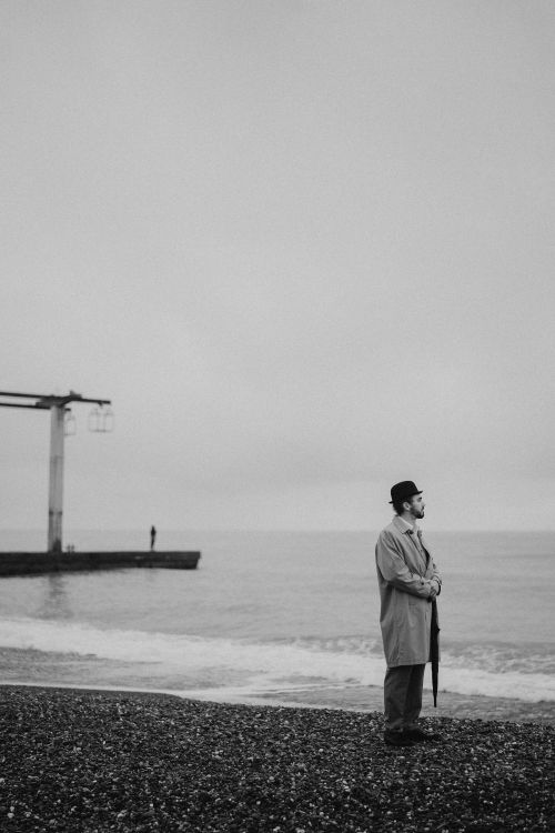 Free A man in a coat stands on a pebble beach in a serene black and white setting. Stock Photo