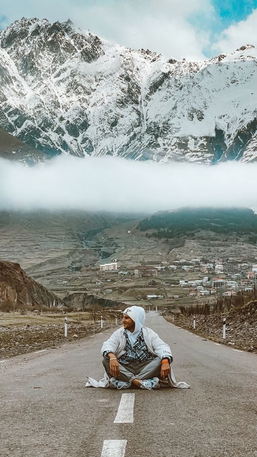 Free A man sits on an empty mountain road with snowy peaks and valley views, evoking travel and adventure. Stock Photo