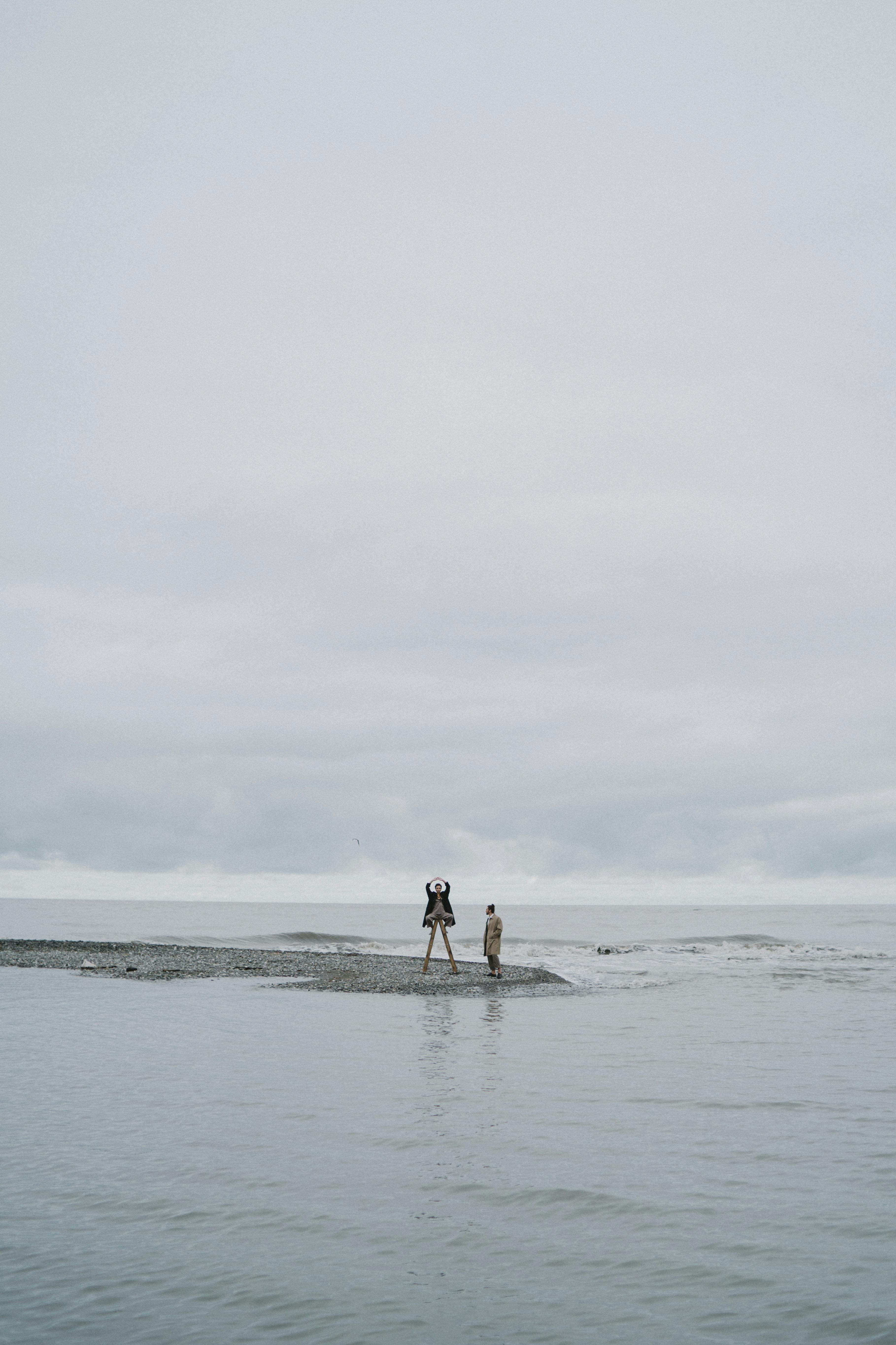 Two people stand on a small sandy beach against a gloomy, overcast seaside backdrop.
