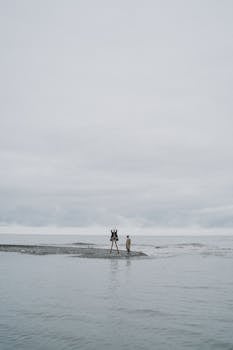 Two people stand on a small sandy beach against a gloomy, overcast seaside backdrop.