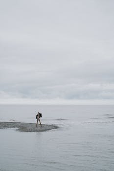 A solitary figure walks on a secluded pebble shore under an overcast sky by the ocean.
