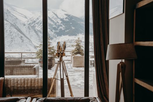 A cozy room with a telescope facing snow-covered mountains through large windows.