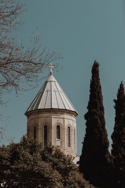 Free A serene church tower stands against a clear blue sky flanked by tall trees, symbolizing peace and heritage. Stock Photo