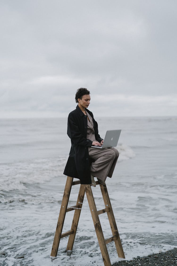 A Woman Sitting On A Wooden Ladder At The Beach While Using Her Laptop