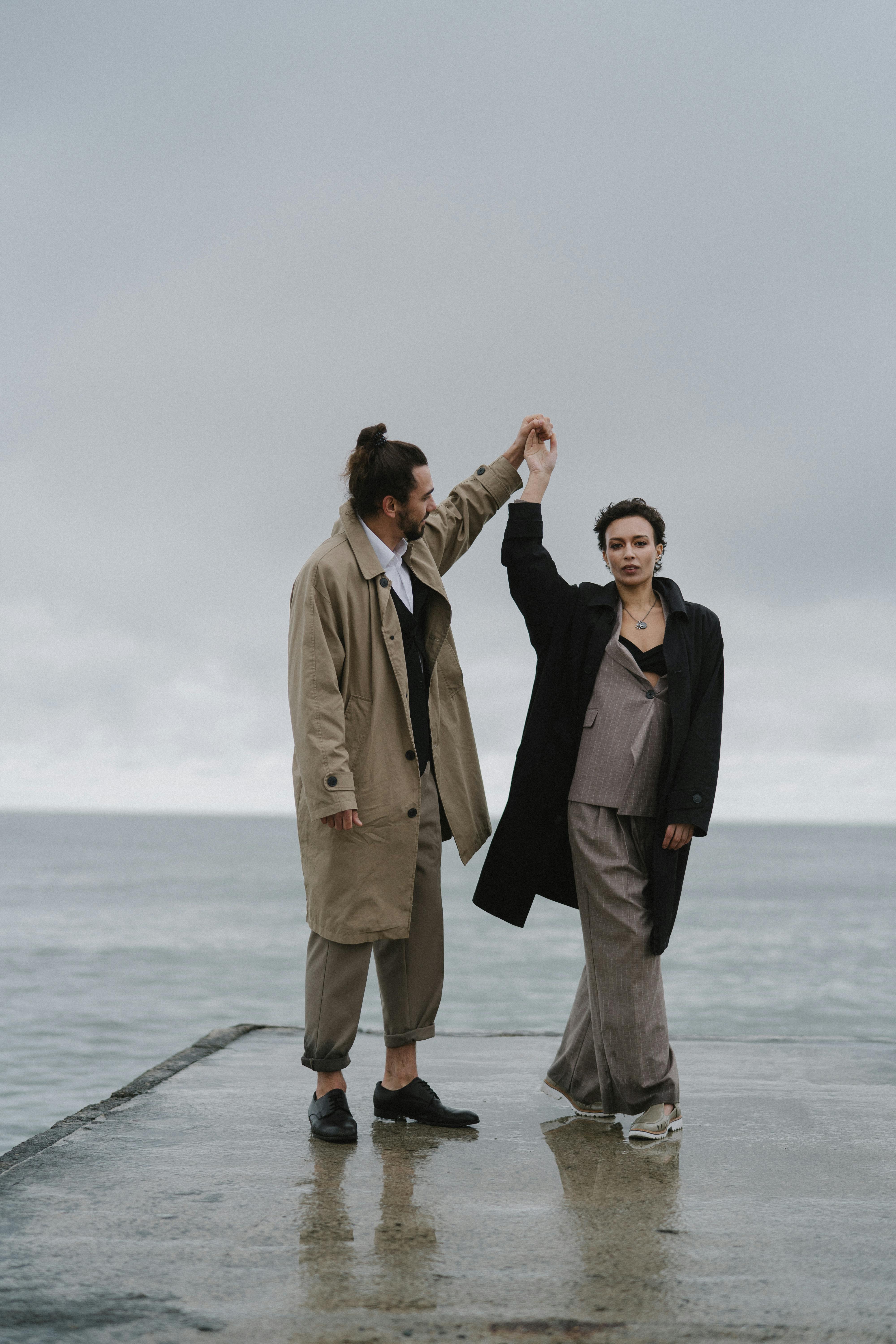 一对优雅的夫妇穿着风衣在海边的码头上跳舞，散发着浪漫和风格