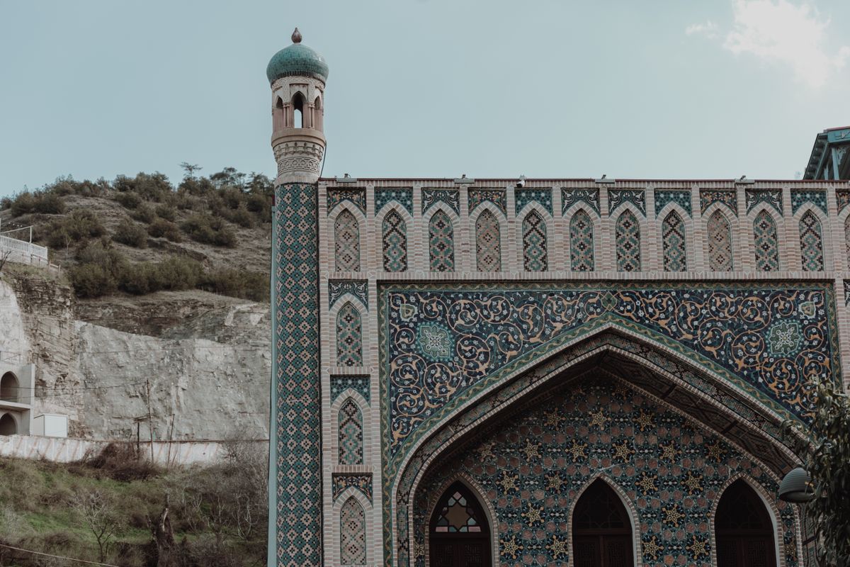 The blue-tiled facade of a bathhouse with a tall tiled turret with a green cap and a large pointed arch with star-pattern tiles, a dry hillside behind